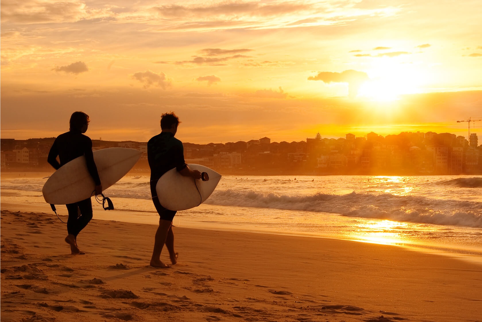 Two Seabourn surfers walk toward the water in the sunset.