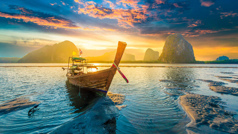 A small speedboat rests by the shore in front of a colorful sunset with hues of yellow, pink, green and blue.